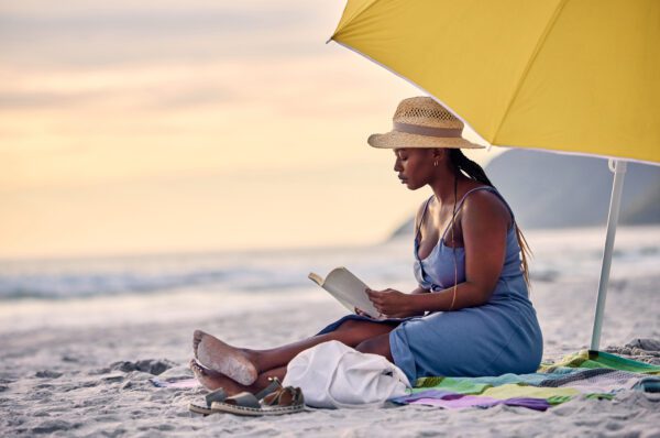 A woman sat on a beach reading underneath a yellow umbrella