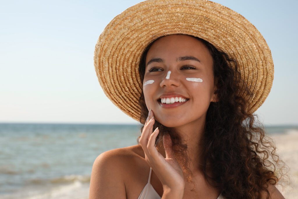 A woman with brown curly hair, wearing a sun hat, smiles at the beach with a stripe of sunscreen on her nose and cheeks