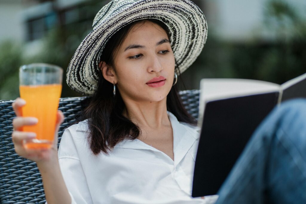 A woman on a sun lounger in a hat holding an orange drink reading a book
