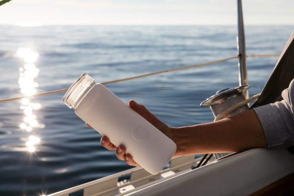 Someone on a boat holds a pink resusable water bottle with a view out to sea in the background.