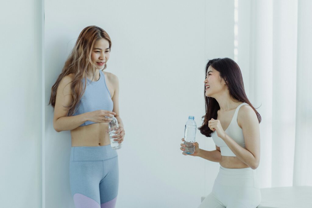 Two women in active wear drink bottles of water indoors while smiling at each other.