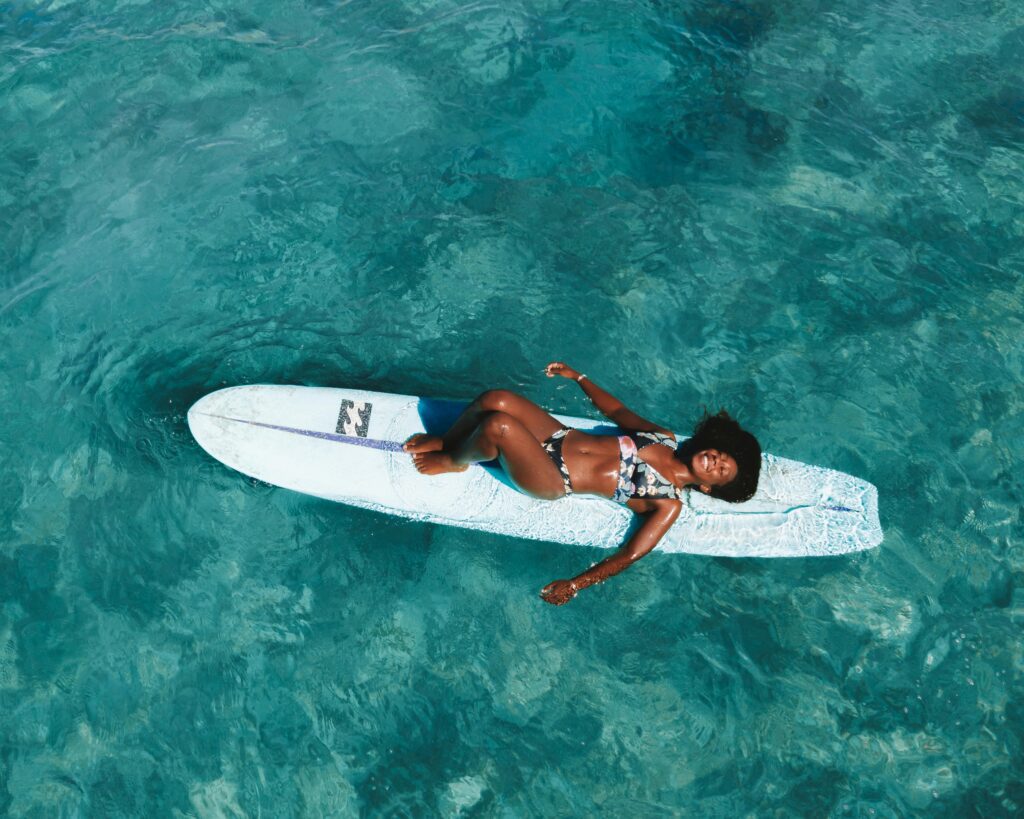 A smiling black woman lies on a surfboard in the ocean.